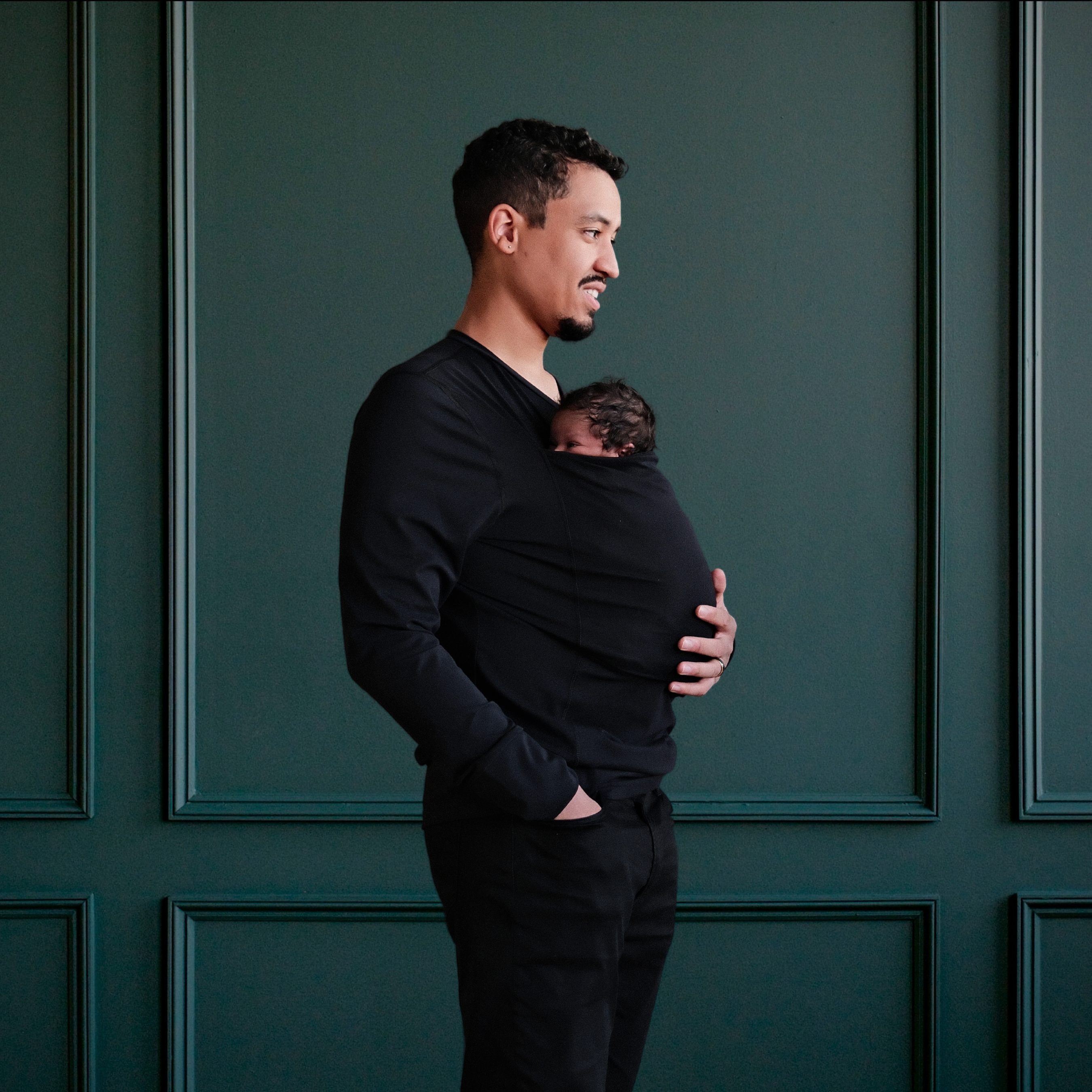 Dad wearing a newborn in a long sleeved black dad shirt in front of a green backdrop.