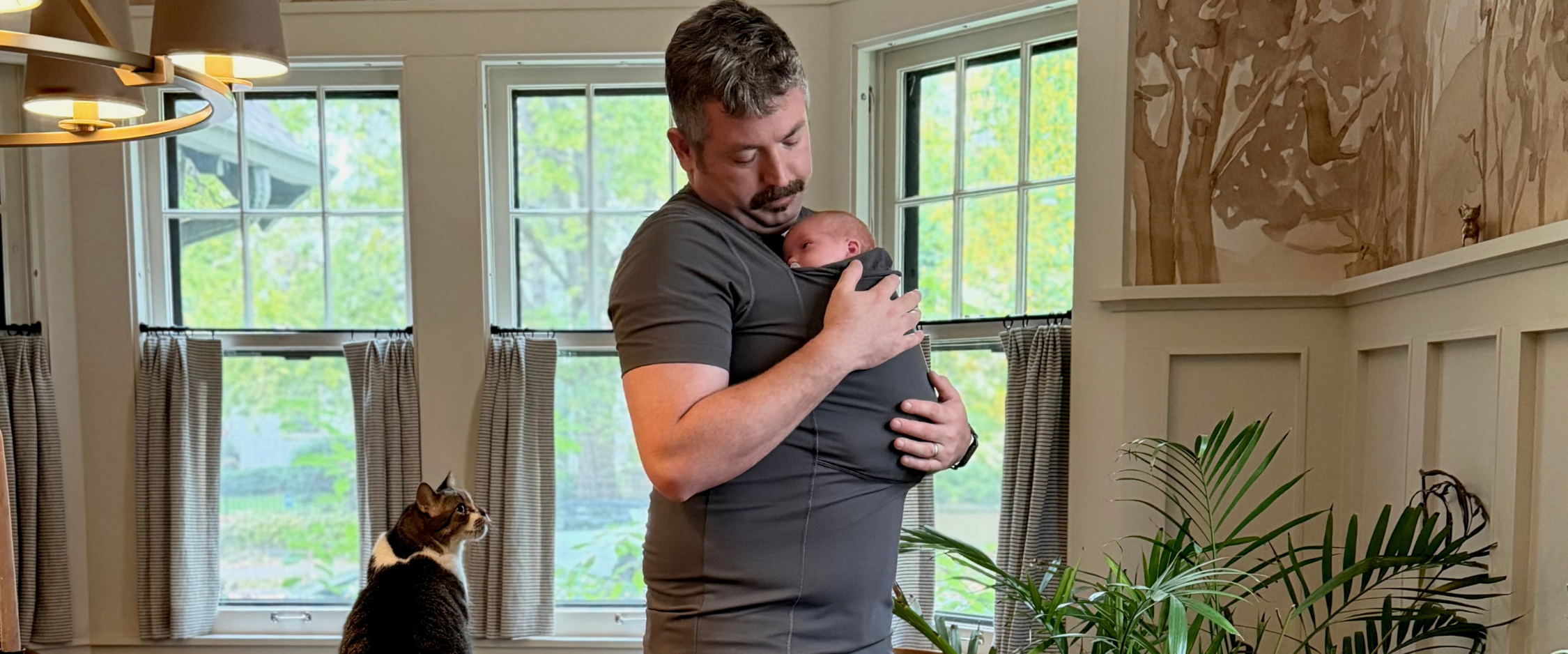 Dad wearing his newborn in the pouch of his Gray Dad Shirt while his cat looks on.