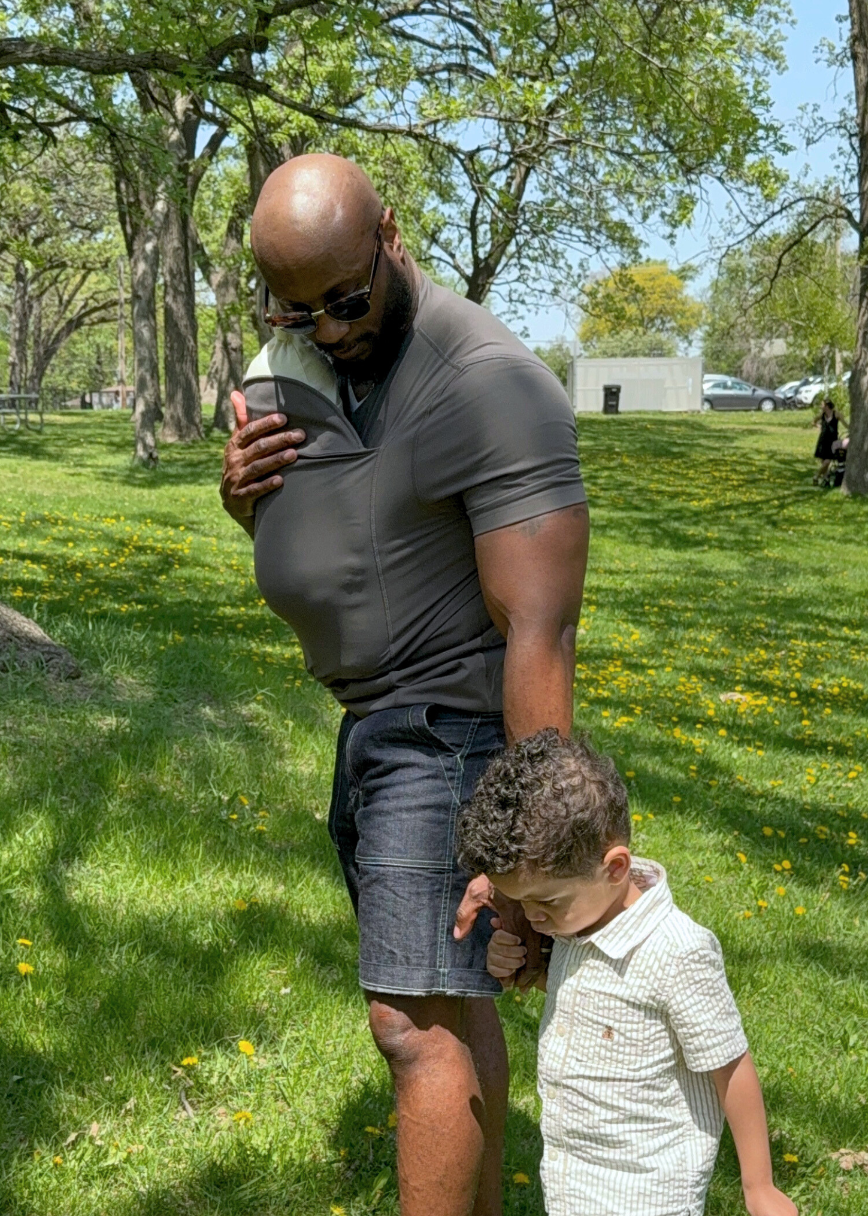 Dad supporting the head of the newborn his dad shirt pouch, while holding hands with his older kid.