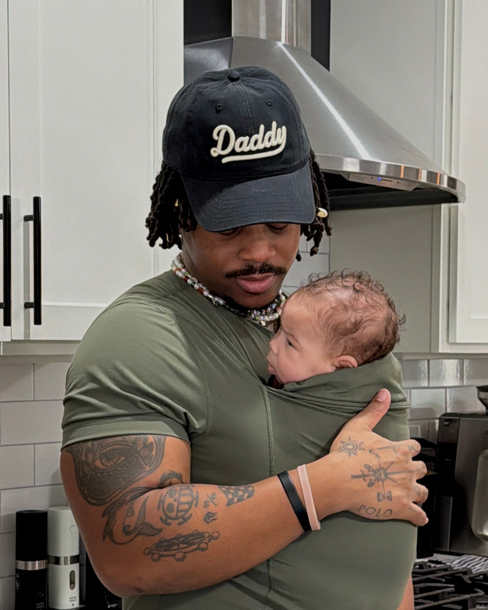 Dad wears his content baby in the pouch of a Fern Dad Shirt, also sporting a black Daddy hat.