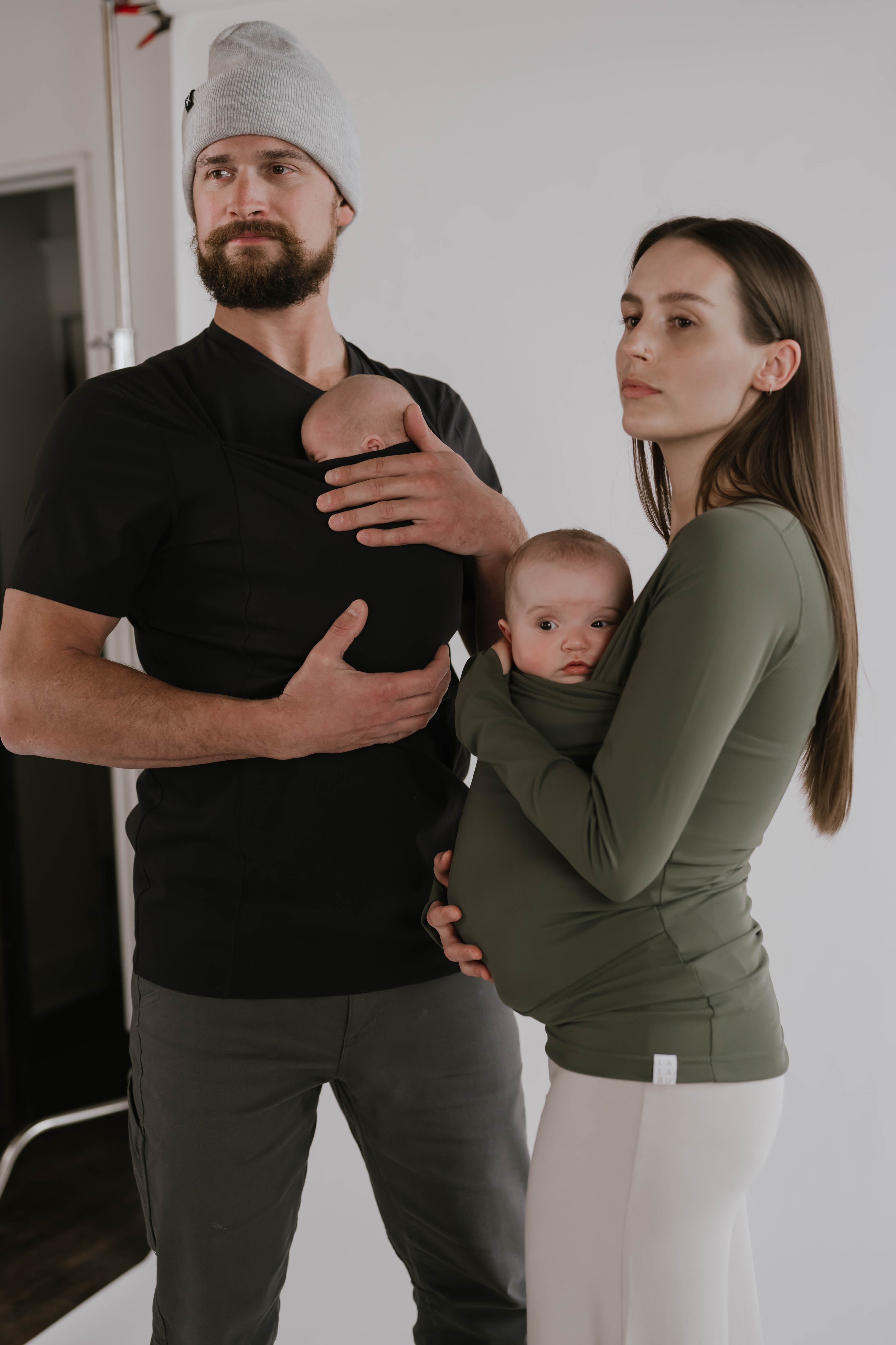 Male wearing a newborn in a black Dad Shirt standing next to a female wearing a newborn in a Fern long sleeve Soothe Shirt.
