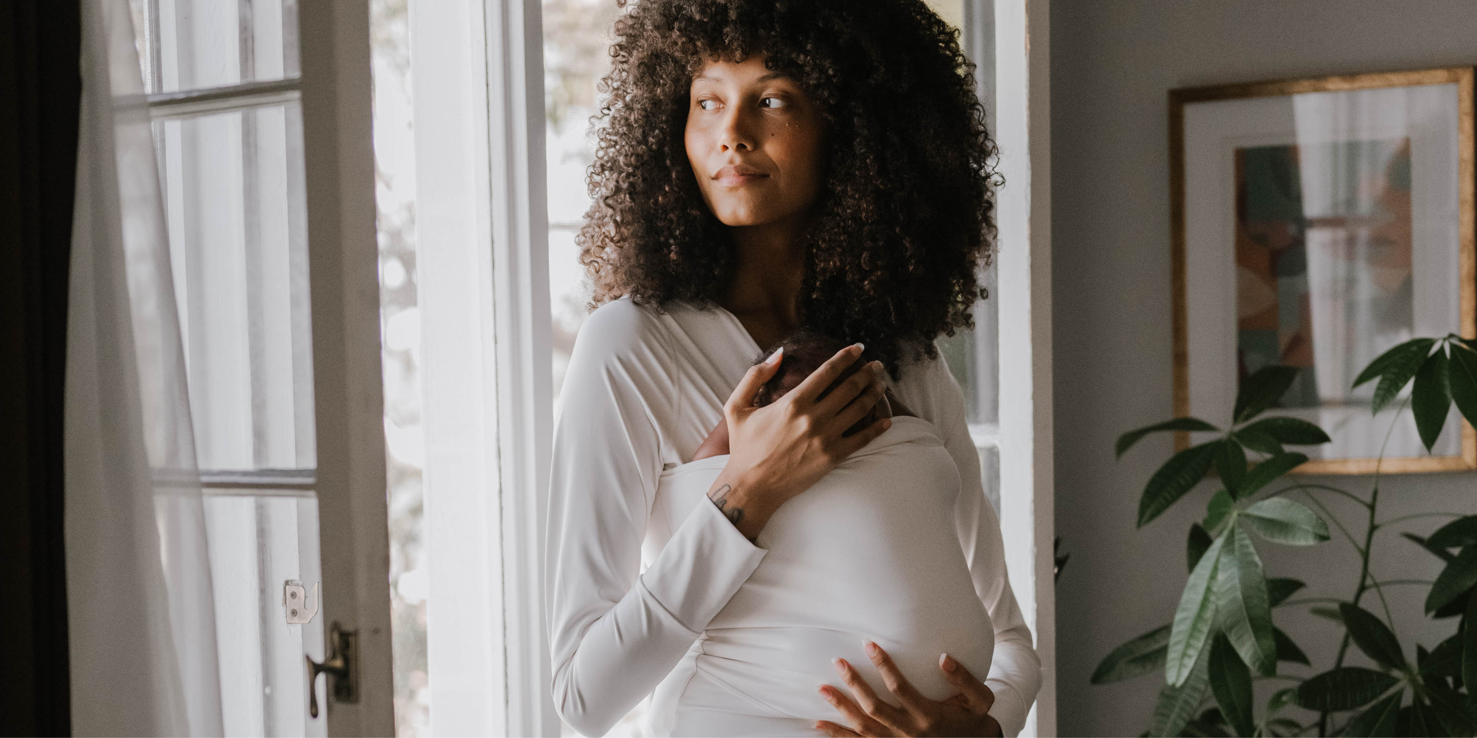 Female in front of opened doors wearing a newborn in a Natural long sleeve Soothe Shirt.