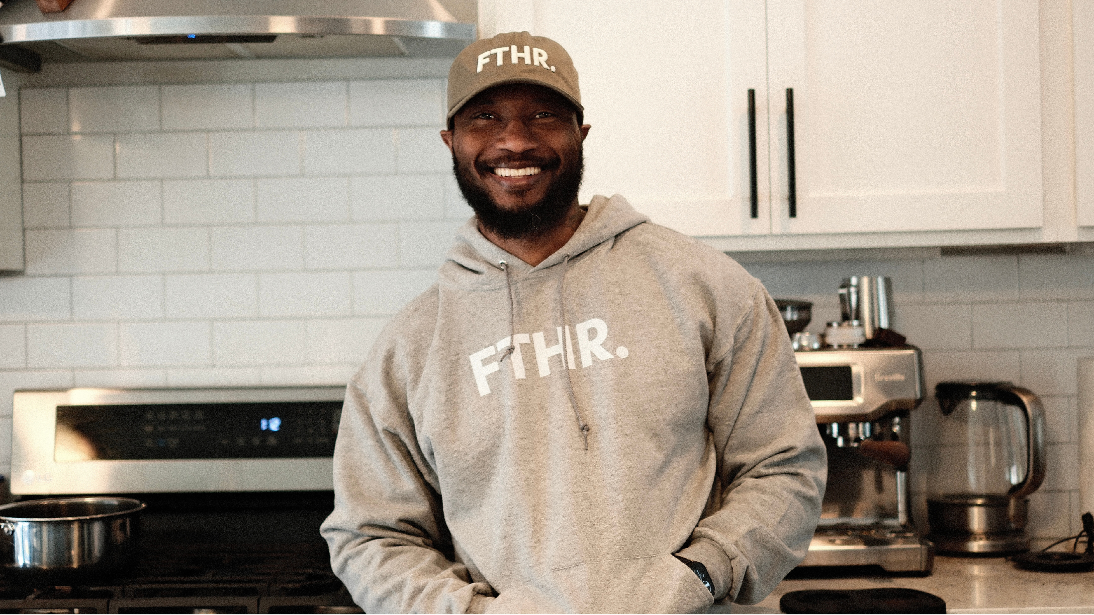 Dad wearing the Lalabu 'FTHR.' green cap and gray hoodie in a kitchen.