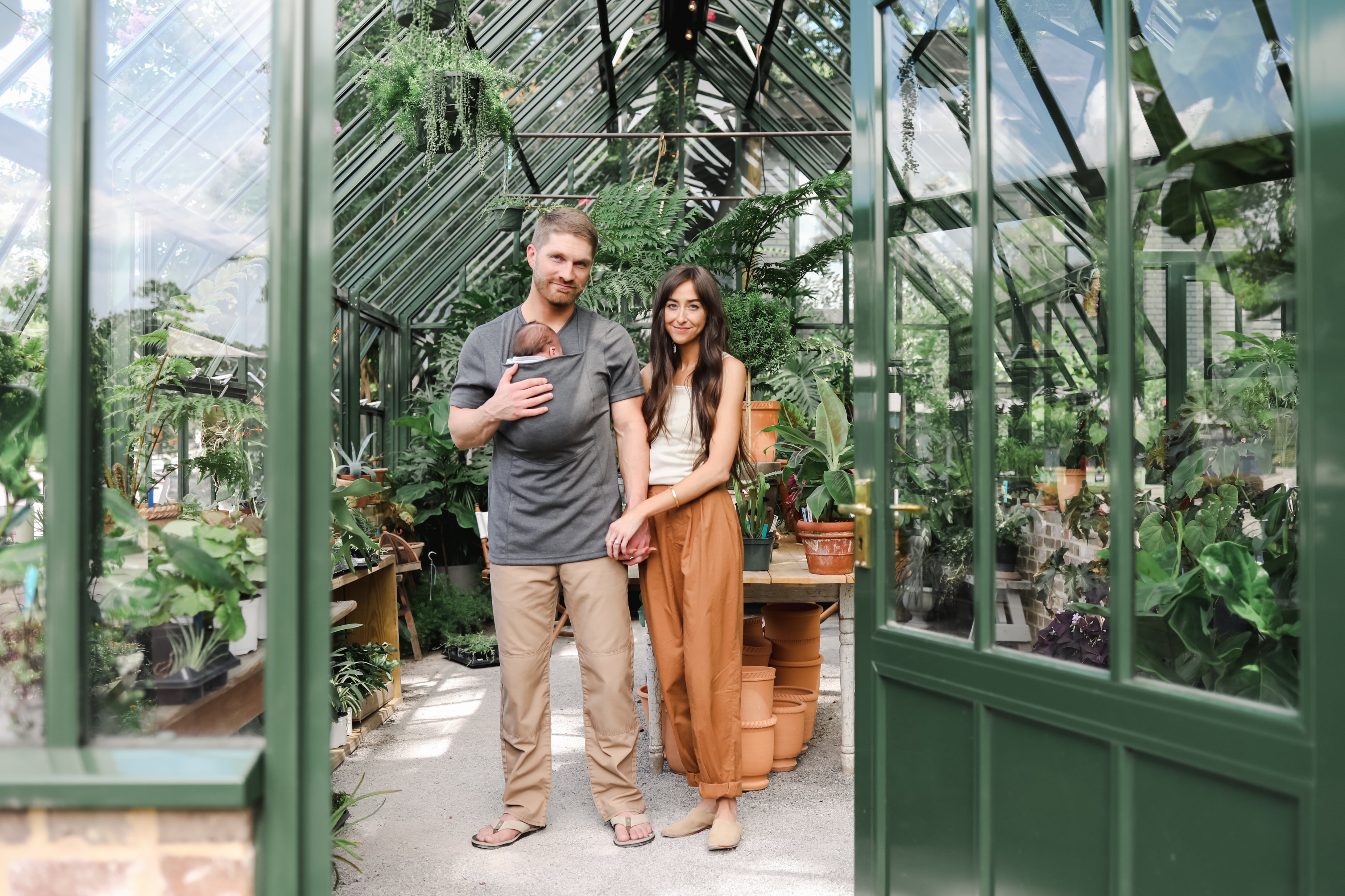 A man and a woman standing inside a greenhouse surrounded by plants, with the man wearing a baby in Dad Shirt baby carrier.