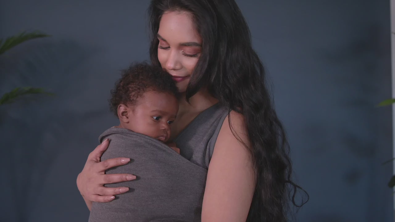 Load video: Female in a grey Soothe Shirt demonstrates how to open the pouch of the shirt, hold the newborn over her shoulder while gently lowering a newborn into it, feet first, and then gently tugging the pouch fabric up with both hands to ensure a secure fit for the baby. The Lalabu logo is shown in white over a navy background at the end.