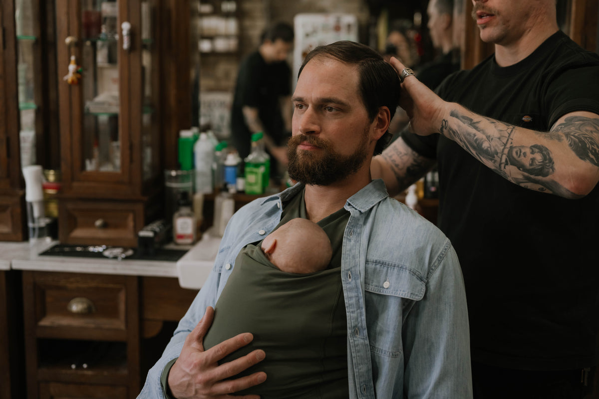 Male wearing a newborn in a Fern Dad Shirt while receiving a haircut.