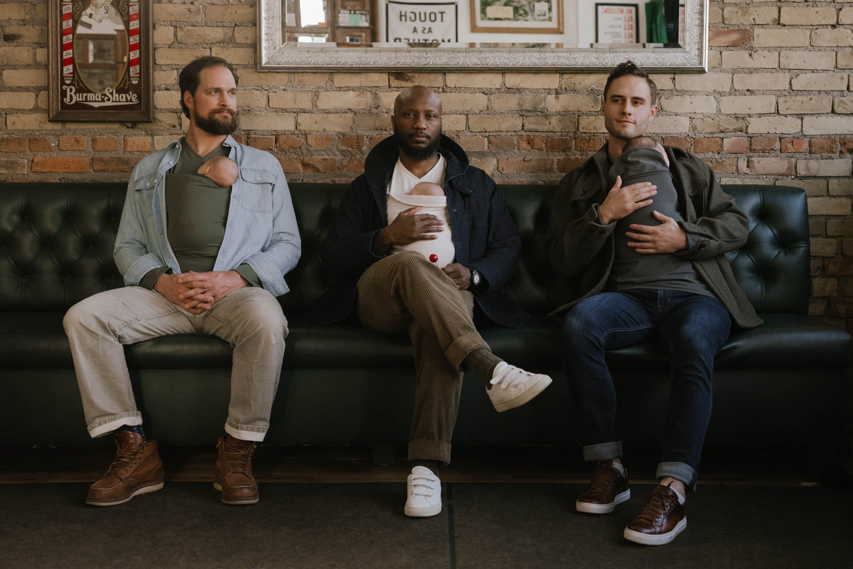 3 males sitting on a barbershop couch, each wearing a newborn in a Dad Shirt.
