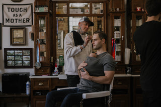 Male wearing a newborn in a Dad Shirt, receiving a hair cut from a male Barber.