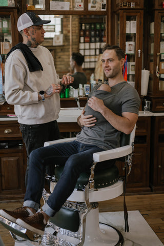 Male sitting in a barber's chair wearing a newborn in a gray Dad Shirt, consulting with a male Barber.