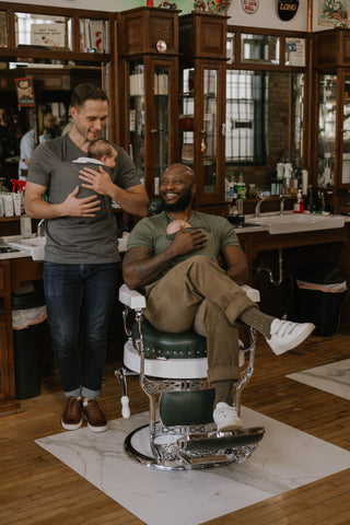 Two males wearing newborns in Dad shirts, one sitting in a barber's chair, the other leaning against a sink.