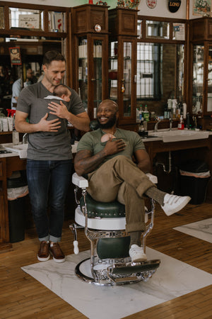 2 males wearing babies in Dad shirts, while sitting in a barber