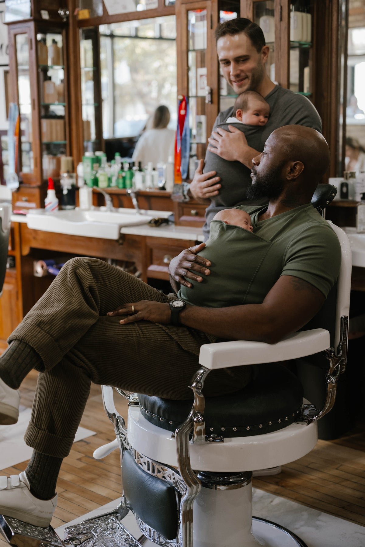 2 males in a barbershop, one sitting wearing , one standing both are wearing newborns in Dad Shirts.