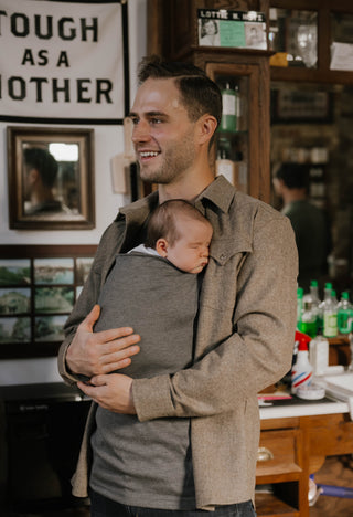 Male wearing a newborn in a gray Dad Shirt in a barbershop.
