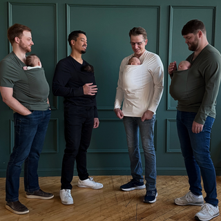 Four dads wearing their newborns in Dad shirts in front of a green backdrop.