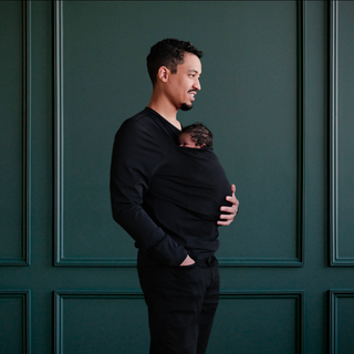 Dad wearing a newborn in a long sleeved black dad shirt in front of a green backdrop.