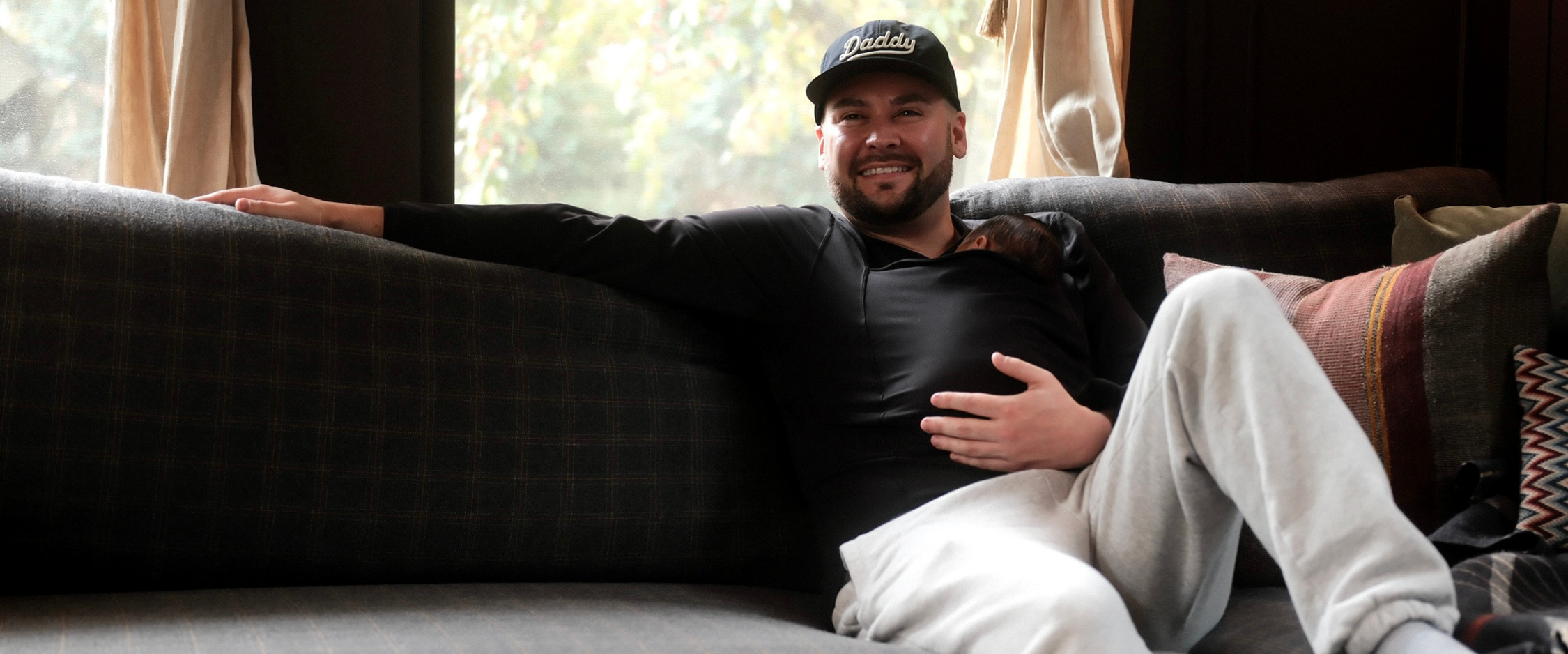 Dad relaxing on a couch wearing his baby in a Black long sleeve Dad Shirt, sporting a Daddy hat.