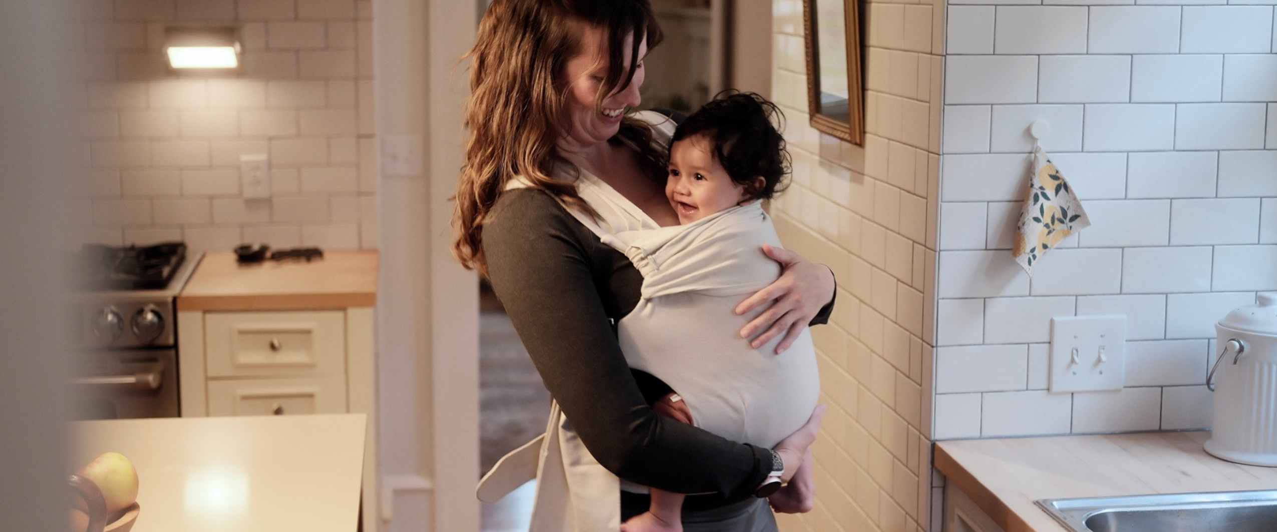 Mom smiling at the baby she's front carrying in a Fog Simple Wrap in the kitchen.