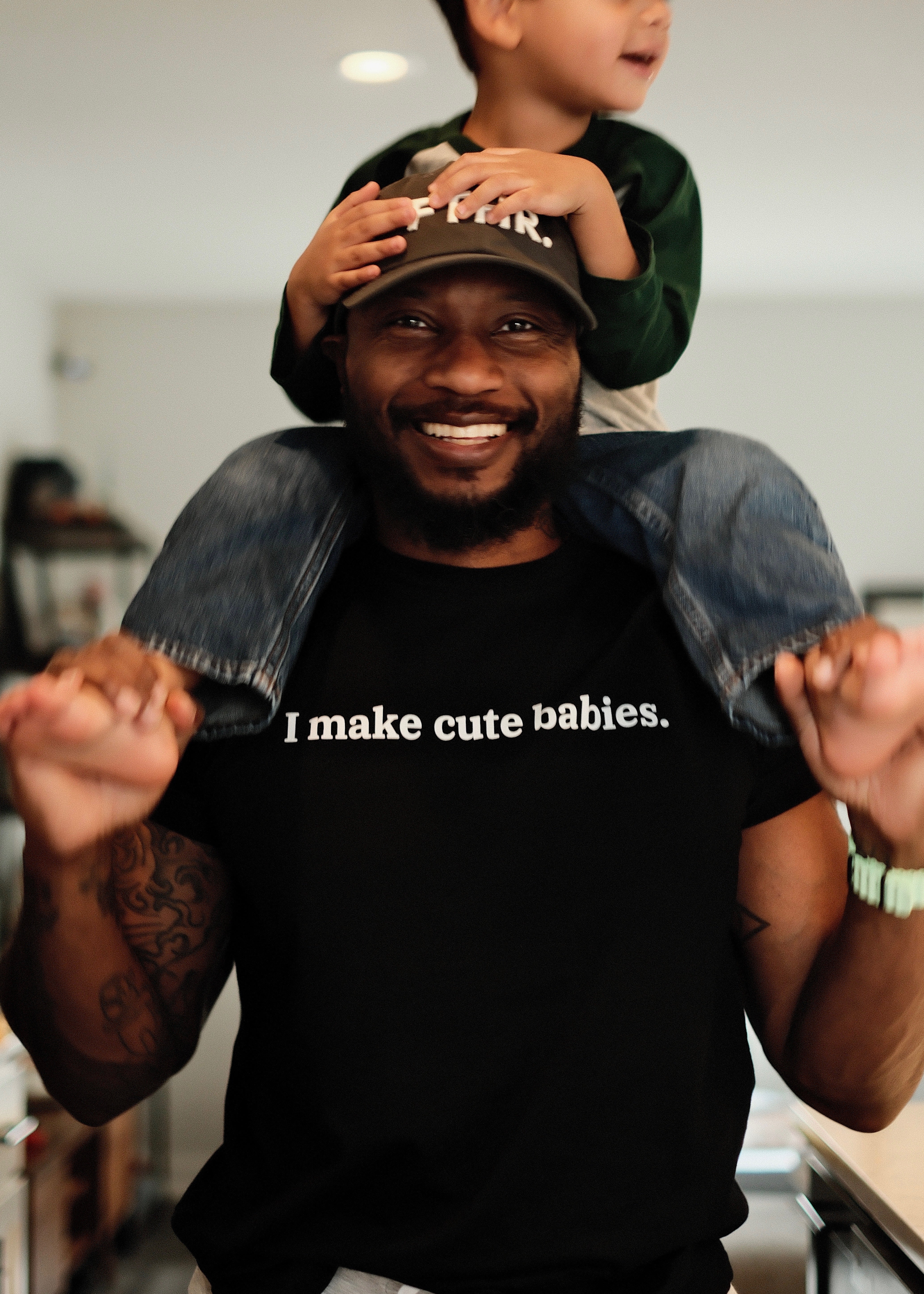 Dad wearing a black t-shirt with 'I make cute babies.' text, holding a child on his shoulders.