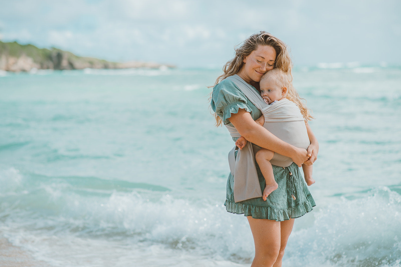 Female front carrying a baby in a Flax Simple Wrap in front of the ocean.