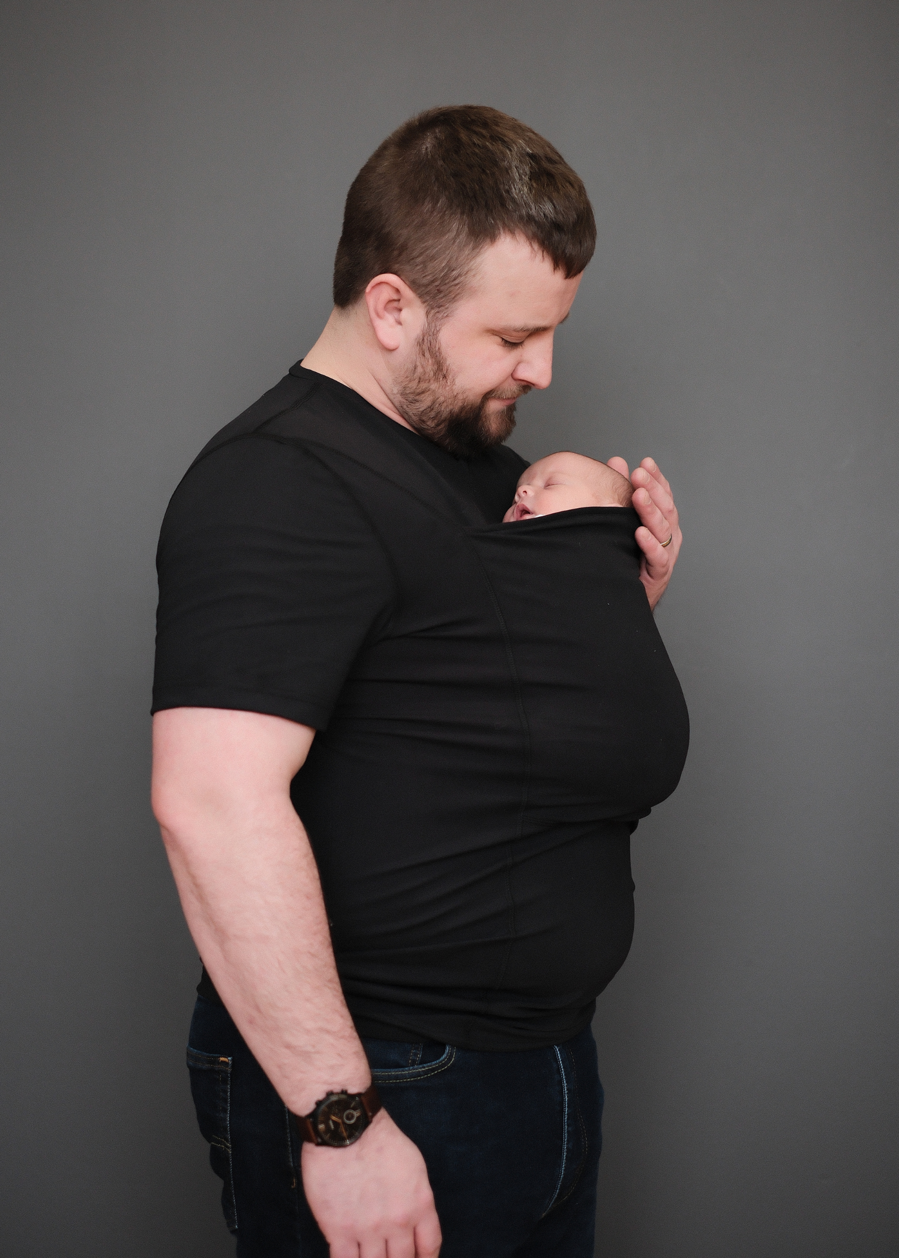 Side profile of dad wearing his newborn in the pouch of a Black Dad Shirt. 