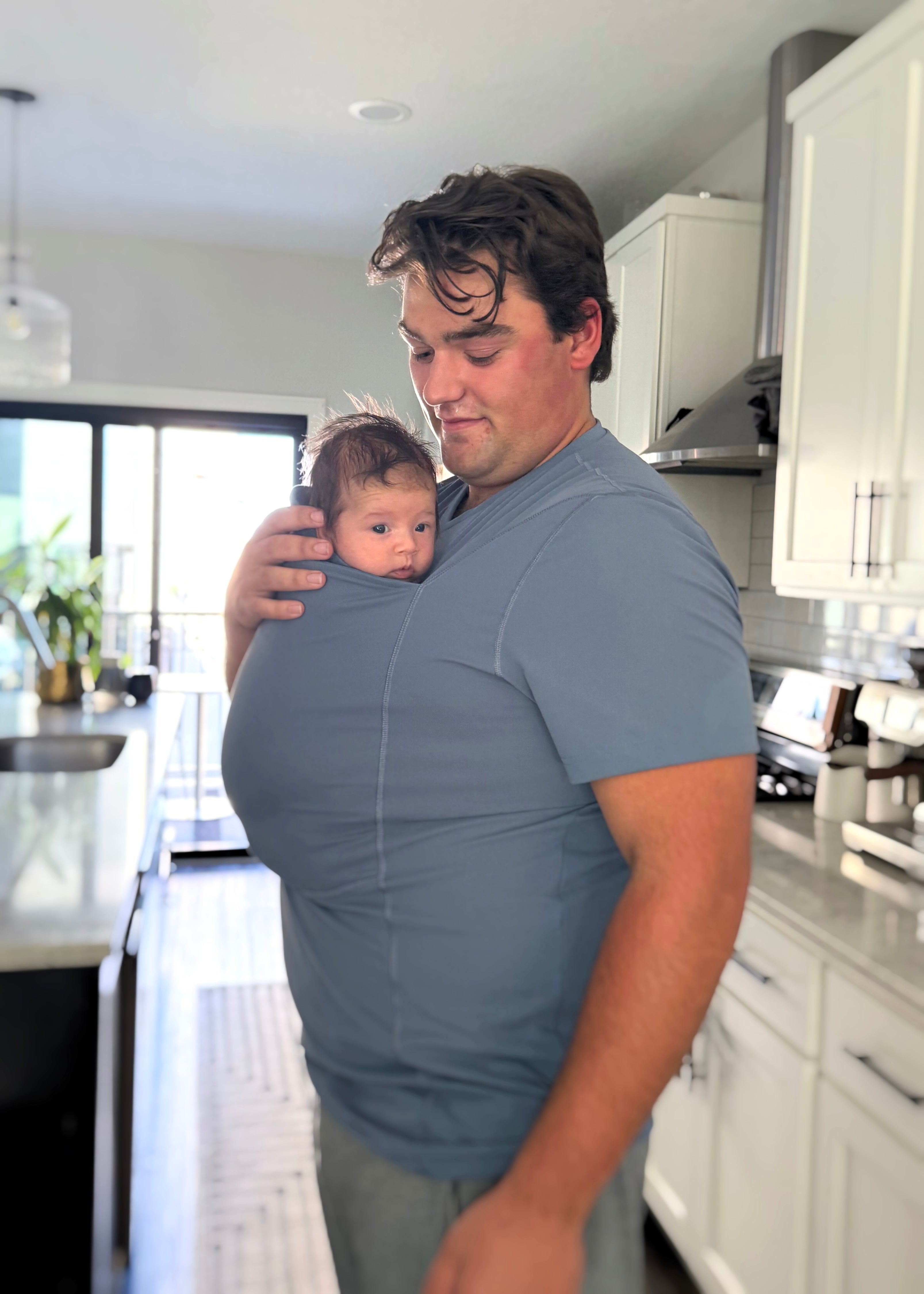 Dad looking down at the baby in the pouch of his Brook Dad Shirt. They are in the kitchen.