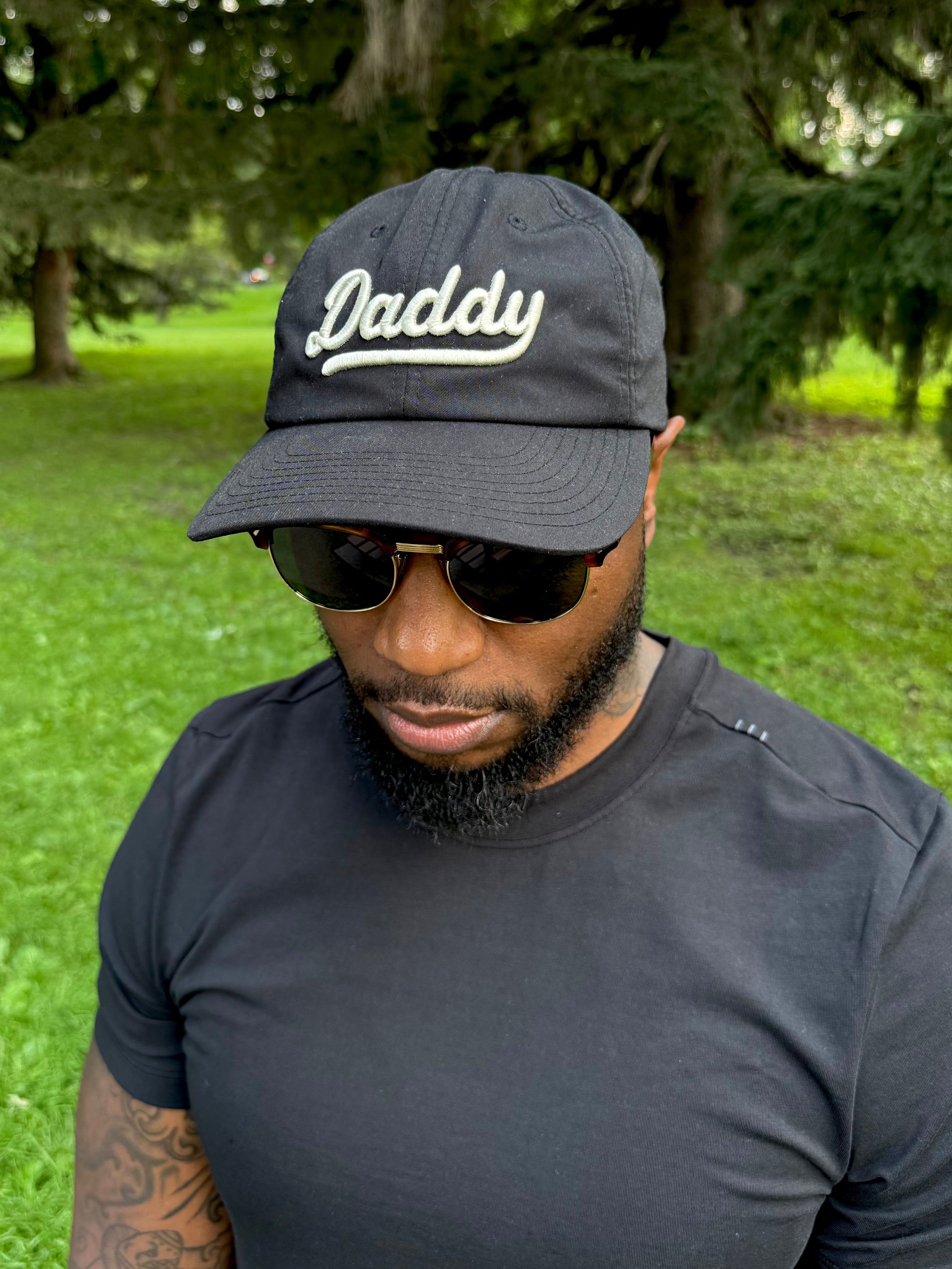 Male showing off his "Daddy" hat, wearing sunglasses and at a park.
