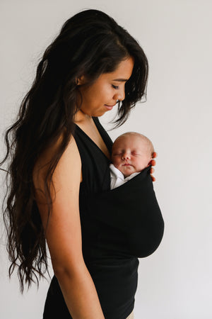 Mom wearing a newborn in a Black Soothe Shirt with a white background.