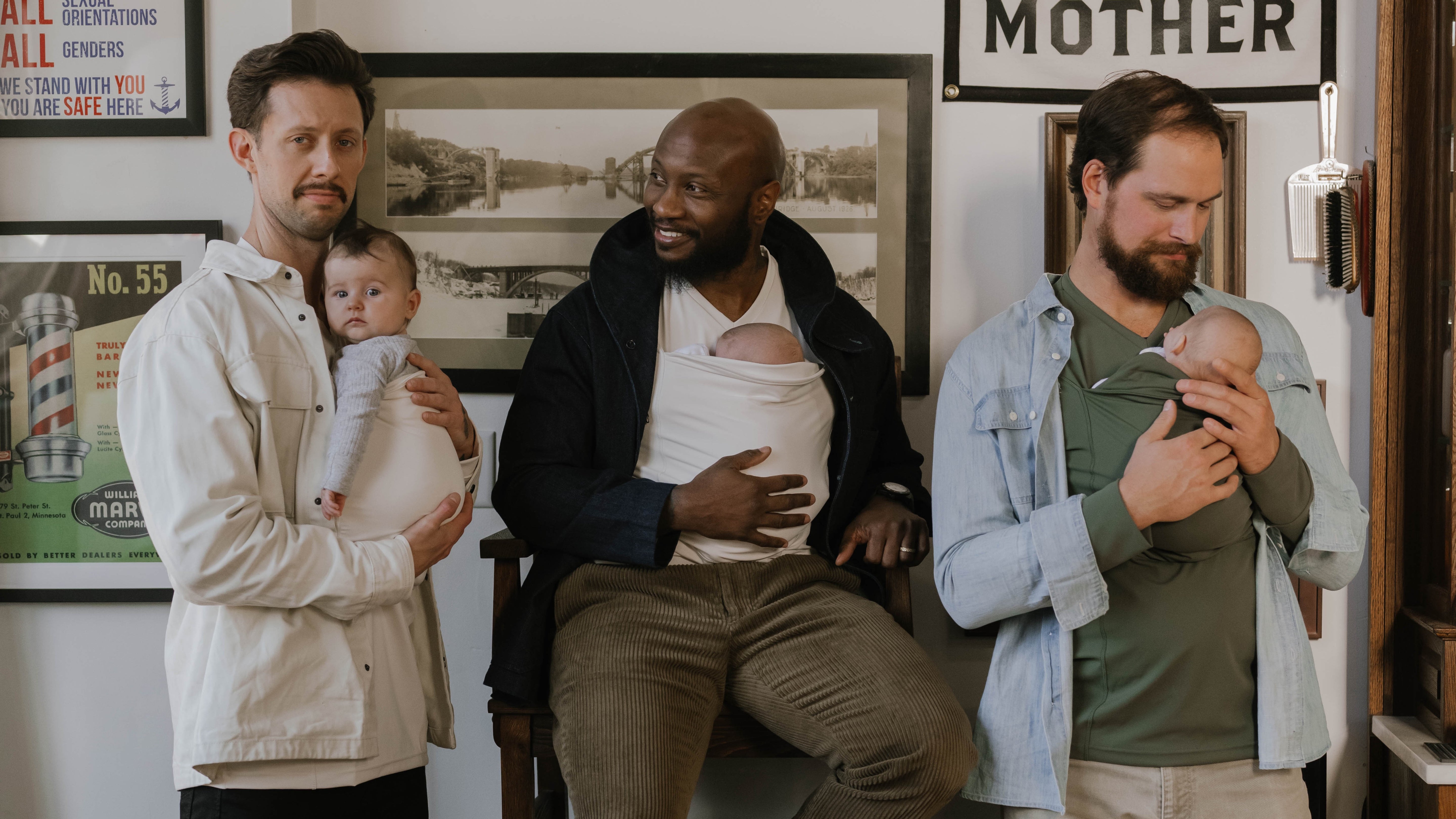 3 Dads wearing newborns in the pouches of their Dad Shirts, in a barbershop.