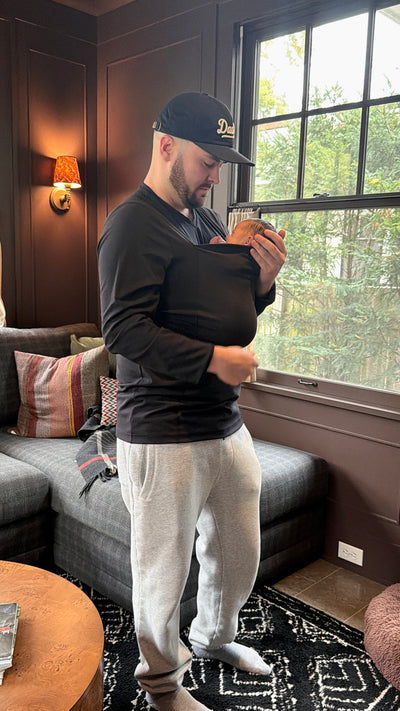 Dad snuggling the newborn in the pouch of his Black long sleeve Dad Shirt sporting a Daddy cap.