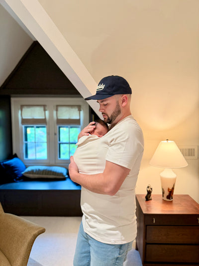 Dad snuggling the sleeping baby inside the pouch of his Natural Dad Shirt in his home.