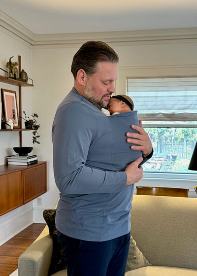 Dad wearing his newborn in a long sleeve Brook Dad Shirt in his living room.