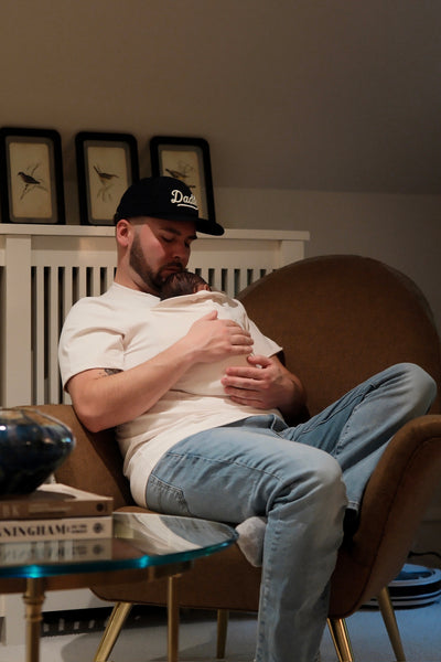Dad snuggling the sleeping baby inside the pouch of his Natural Dad Shirt relaxing in a chair.