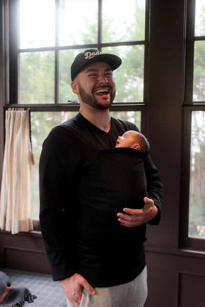 Dad laughing while wearing his newborn in his Black long sleeve Dad Shirt sporting a Daddy cap.