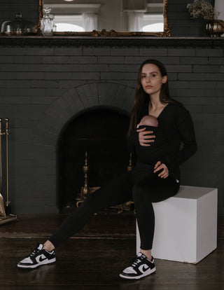 Female sitting on a box, wearing a newborn in a black long sleeve Soothe Shirt.