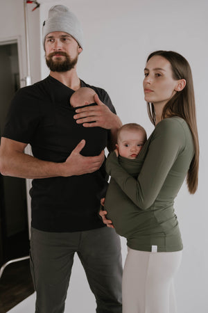Male wearing a newborn in a black Dad Shirt standing next to a female wearing a newborn in a Fern long sleeve Soothe Shirt.