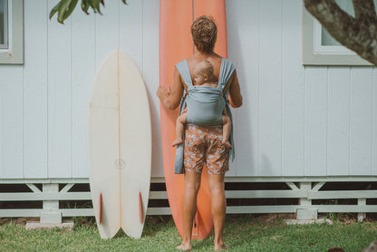 Dad leaning a surfboard against the house while back carrying baby.