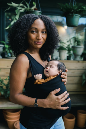 Mom wearing a newborn in a black Soothe Shirt inside a greenhouse.