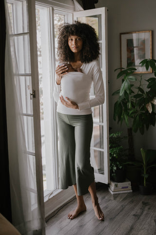 Full body view of a female in front of opened doors wearing a newborn in a Natural long sleeve Soothe Shirt.