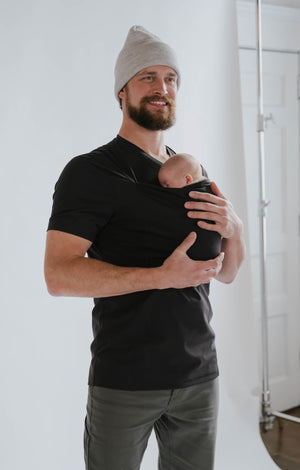 Dad wearing a newborn in a black Dad Shirt with a white backdrop.