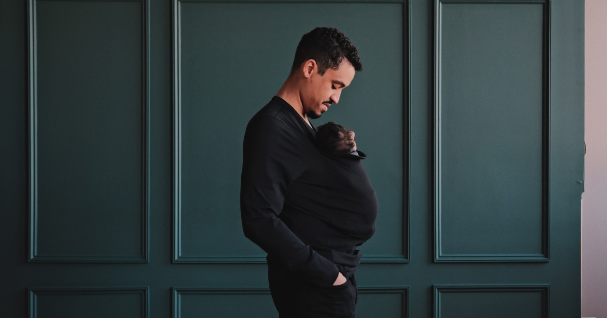 Dad wearing his newborn in a black long sleeved Dad Shirt in front of a green backdrop.