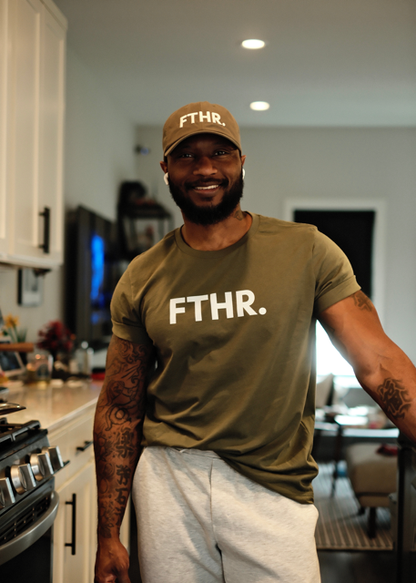 Dad wearing the Lalabu green 'FTHR.' t-shirt and cap in a kitchen.