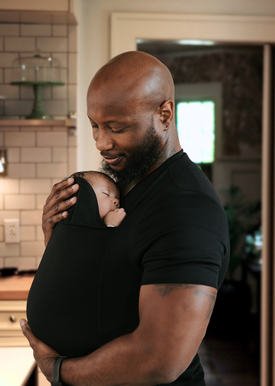 Dad snuggling the sleeping baby inside the pouch of his Black Dad Shirt in a kitchen.