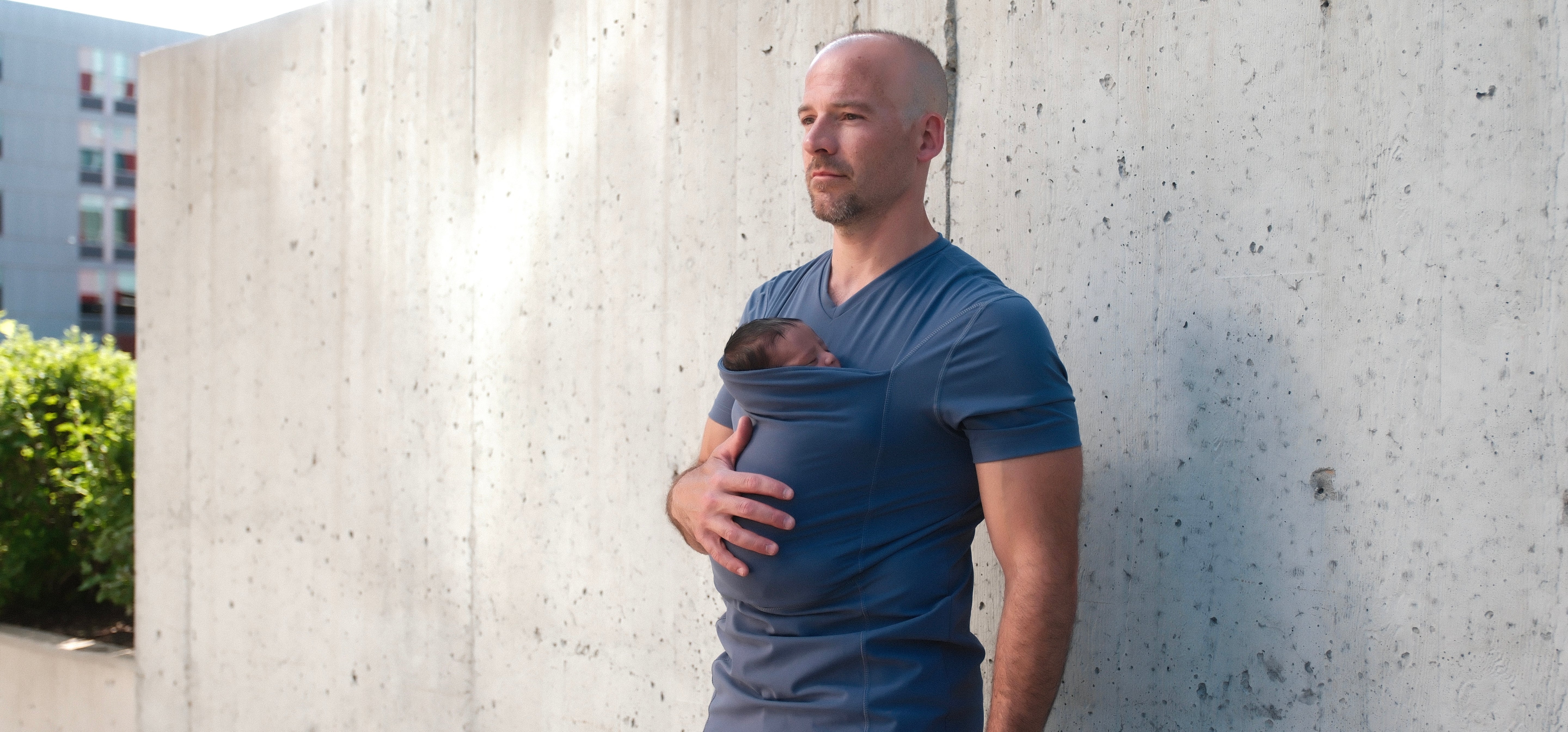 Dad wearing his baby in the pouch of a Brook Dad Shirt in front of a concrete wall.