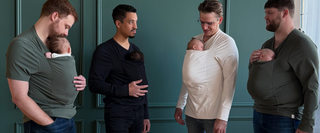 Four dads wearing their newborns in Dad shirts in front of a green backdrop.