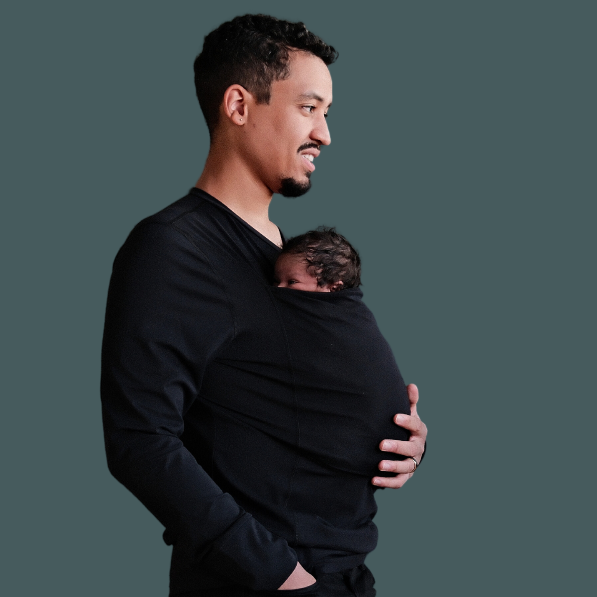 Dad wearing a newborn in a black long sleeved dad shirt in front of a green background.