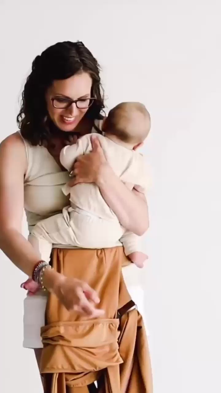 Mom demonstrates putting the straps of the Simple Wrap over her and baby's heads, puts her arms through the straps and looks at her baby.