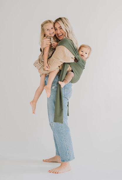 Female back carrying a baby in a Fern Simple Wrap, while holding a child.